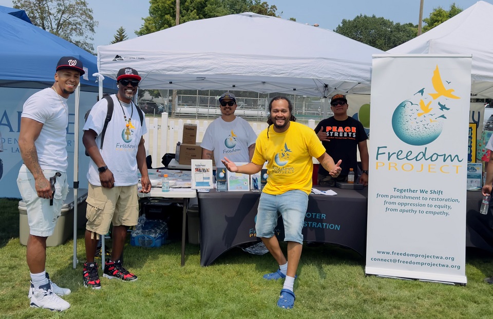 Volunteers staff a Freedom Project booth at an outdoor community event. Four men stand behind tables under white canopies, with organization banners and informational materials visible. The setting appears to be a sunny park or sports field.